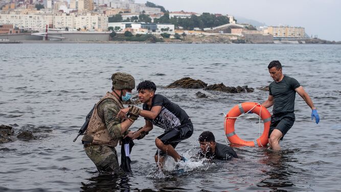 Un soldado del Ejército ayuda a dos migrantes a salir del agua en la playa de El Tarajal.